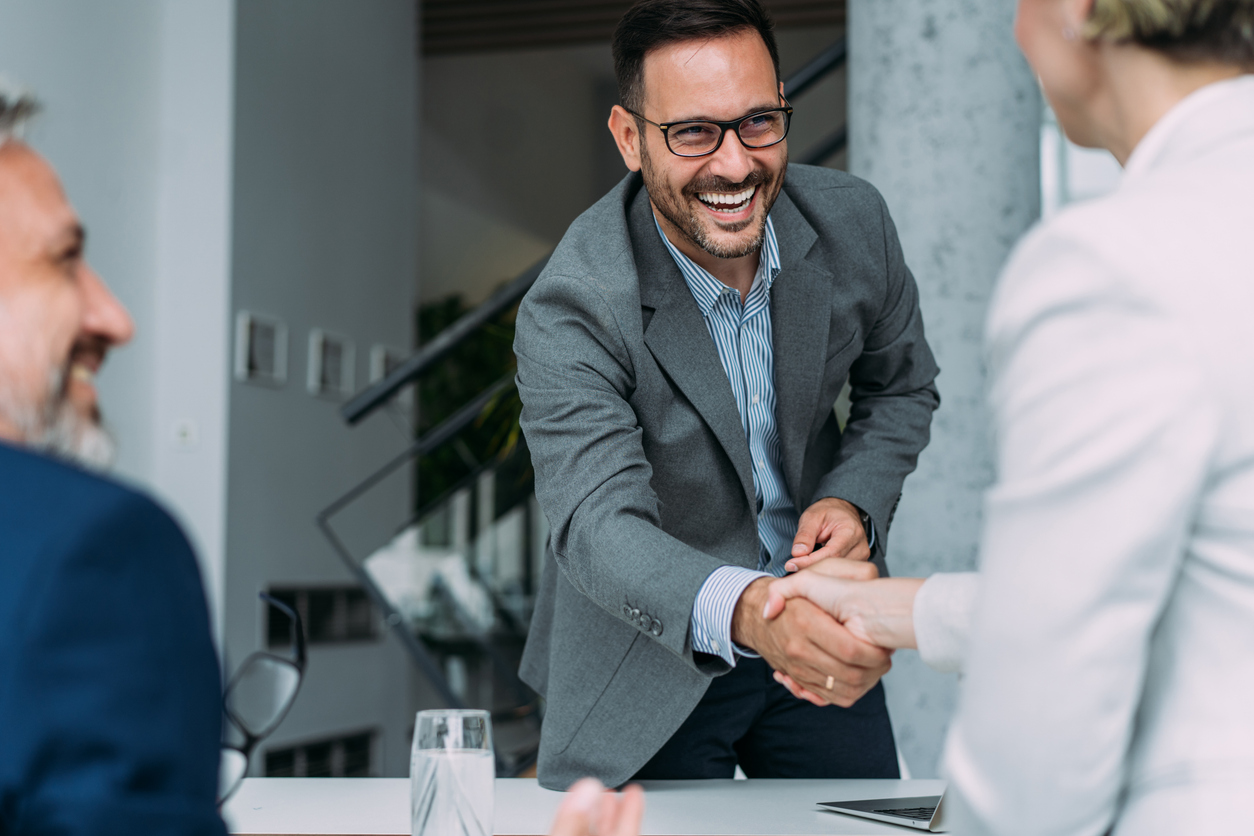 During a meeting in a modern office, business people in suits are shaking hands across the table. In the background, a group of business people are having a meeting. In the foreground, three entrepreneurs are meeting in a board room.